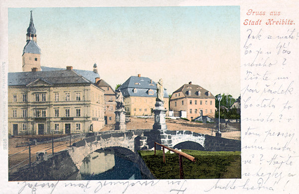 Diese Ansichtskarte vom Anfange des 20. Jahrhunderts zeigt die Nordseite des Marktplatzes vor der St. Georgskirche noch mit der ursprüglichen Steinbrücke über den Chřibská Kamenice-Bach, auf der die Statuen der Immaculata und des hl. Johannes v. Nepomuk von Franz Werner standen.