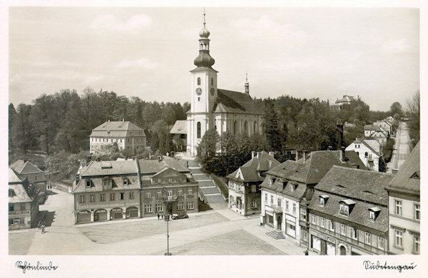 Diese Ansichtskarte zeigt die Nordwestecke des Marktplatzes um das Ende der 30er Jahre des 20. Jahrhunderts. Statt des alten Hauses an der Ecke des Platzes sieht man hier schon den Neubau mit der Apotheke, und die Gaststätte „Zum Rathaus“ ist durch das Gebäude der Handels- und Industriebank verdrängt worden. Das Nachbarhaus hat sich teilweise seinen ursprüngkiche Charakter erhalten, trotzdem sein Laubengang in Geschäfte umgewandelt wurde. Auf der Anhöhe über dem Marktplatz steht die barocke Kirche der hl. Maria Magdalena mit der Pfarre.