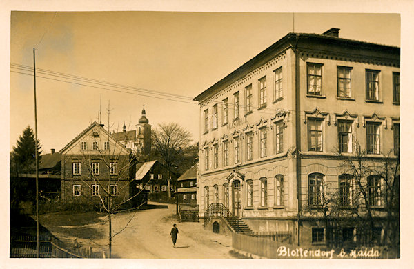 Diese Ansichtskarte zeigt das monumentele Gebäude der Volksschule im Ortszentrum. Im Hintergrund sieht man die Kirche zur Heiligen Dreifaligkeit.