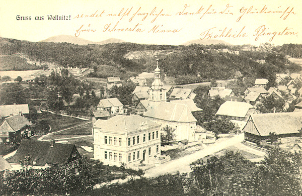 Tato pohlednice z doby před 1. světovou válkou zachycuje střední část Velenic s kostelem Nejsvětější Trojice a patrovou budovou školy z roku 1902, v níž dnes sídlí obecní úřad.