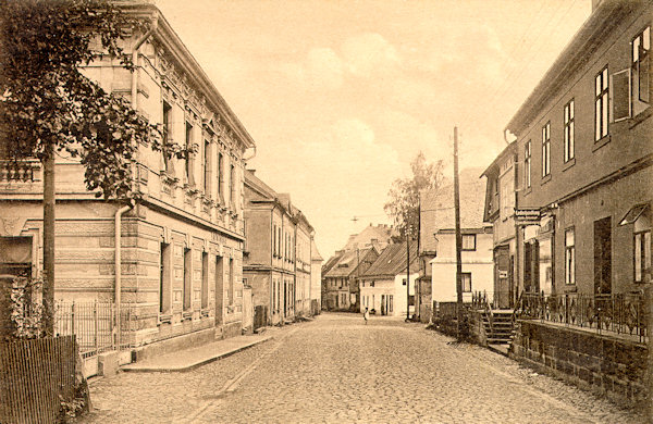 Auf dieser Ansichtskarte sehen wir den unteren Teil der Rumburger Gasse (heute Komenský-Gasse), die in der 30er Jahren nicht mehr dem wachsenden Verkehr entsprach und deshalb wesentlich verbreitert wurde. Dabei wurden fast alle Häuser an der rechten Seite mit Ausnahme die beiden ersten abgerissen.