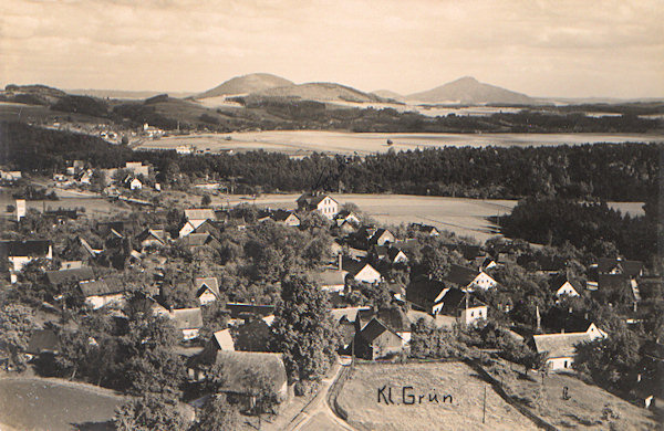 Diese Ansichtskarte aus den 20er Jahren des 20. Jahrhunderts zeigt das Dorf Drnovec (Kleingrün) vom der Stelle am Hang des Zelený vrch (Grünberg) gesehen, an der früher die Gaststätte „Schweizerhaus“ stand. Über dem Dorf erhebt sich der bewaldete Kamm des Hohlsteins, links hinter ihm sieht man die Kirche in Kunratice (Kunnersdorf) und am Horizont ragen die auffallenden Gipfel des Tlustec (Tolzberg), Kovářský vrch (Schmiedsberg) und Ralsko (Roll) empor.