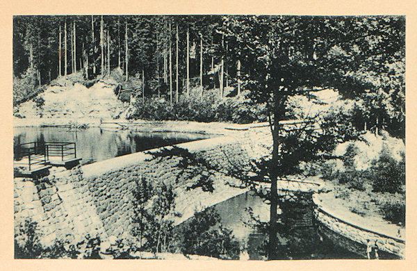 This picture postcard shows the paved damm of the storage reservoir built 1937 to 1938 in the narrow valley of the Hamerský potok-brook. The dam served to drive the mill and the sawmill in the village Hamr situated in the valley down the brook.