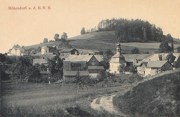 Diese Ansichtskarte zeigt das Ortszentrum mit der im Jahr 1788 erbauten Dreifaltigkeits-Kapelle. Links hinter den Häusern ist der Bahnhof und über ihm sieht man die Kuppe des Sokolík-Hügels (Falkenberg).